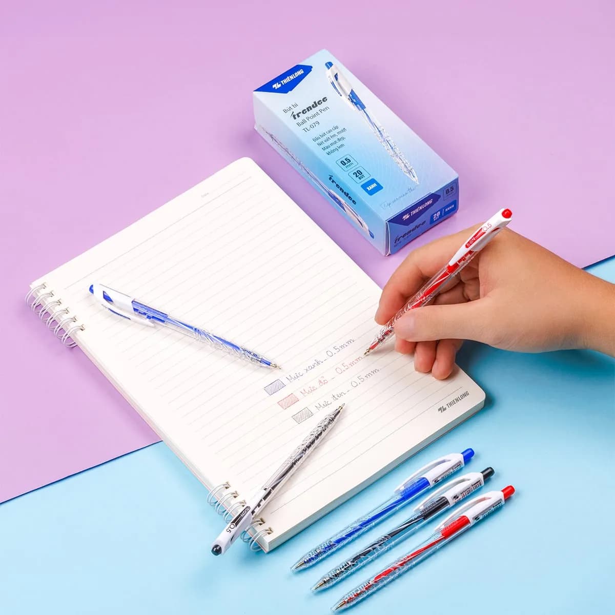 Người dùng đang viết lên sổ bằng bút bi Thiên Long TL-079, bên cạnh là hộp bút và các loại bút mực xanh, đỏ, đen.