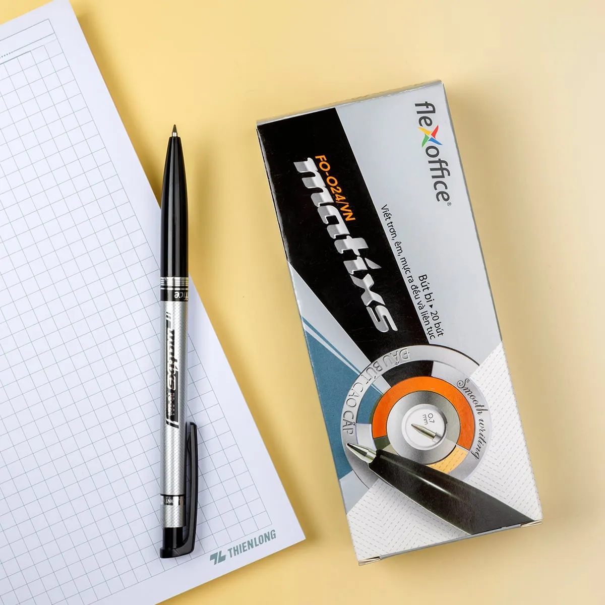 Bút bi FlexOffice FO-024 0.7mm bên cạnh vỏ hộp sản phẩm và cuốn sổ kẻ ô trên nền vàng nhạt