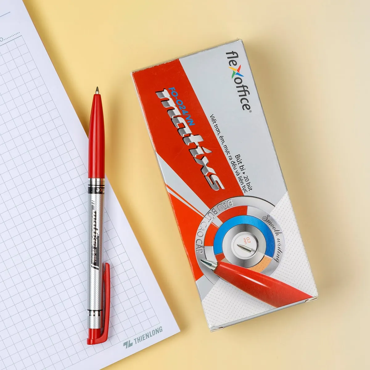 Hộp bút bi Flexoffice FO-024 0.7mm và một chiếc bút đỏ đặt trên nền giấy kẻ ô ly.